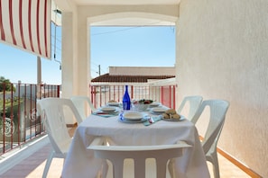 Varanda com mesa posta para refeições, cadeiras de plástico branco e vista para um edifício com telhado de azulejos.