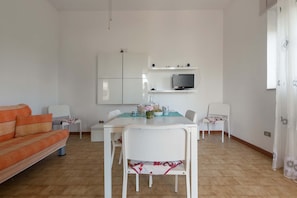 Uma sala de estar moderna com um sofá, uma mesa de jantar com cadeiras, uma TV fixada na parede e uma área de cozinha.