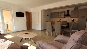 Un salon moderne avec un canapé marron, une table basse ronde et un coin repas, la cuisine étant visible au fond.