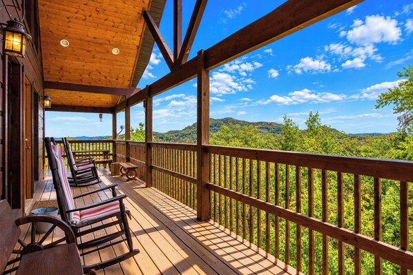 A wooden deck with rocking chairs and tables, overlooking a lush green landscape.