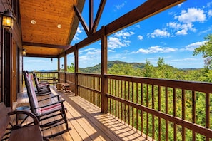 A wooden deck with rocking chairs and tables, overlooking a lush green landscape.