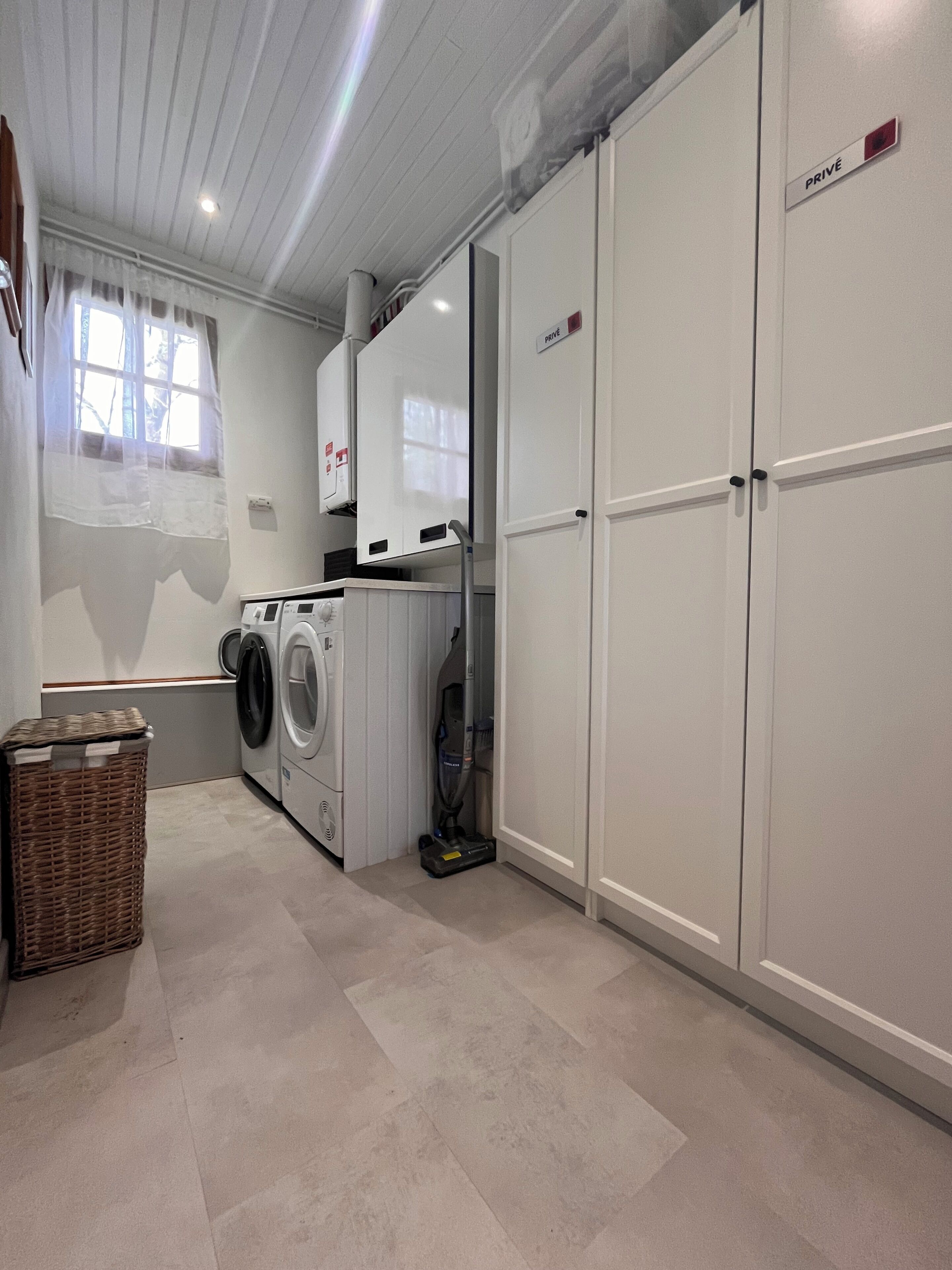 A laundry room with a washing machine, dryer, and vacuum cleaner. There is a wicker basket and a white cabinet with a 'PRIVÉ' sign.