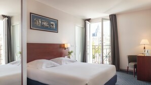 Habitación de hotel con dos camas, un escritorio, una silla, un balcón con vistas a edificios y un cuadro enmarcado en la pared.