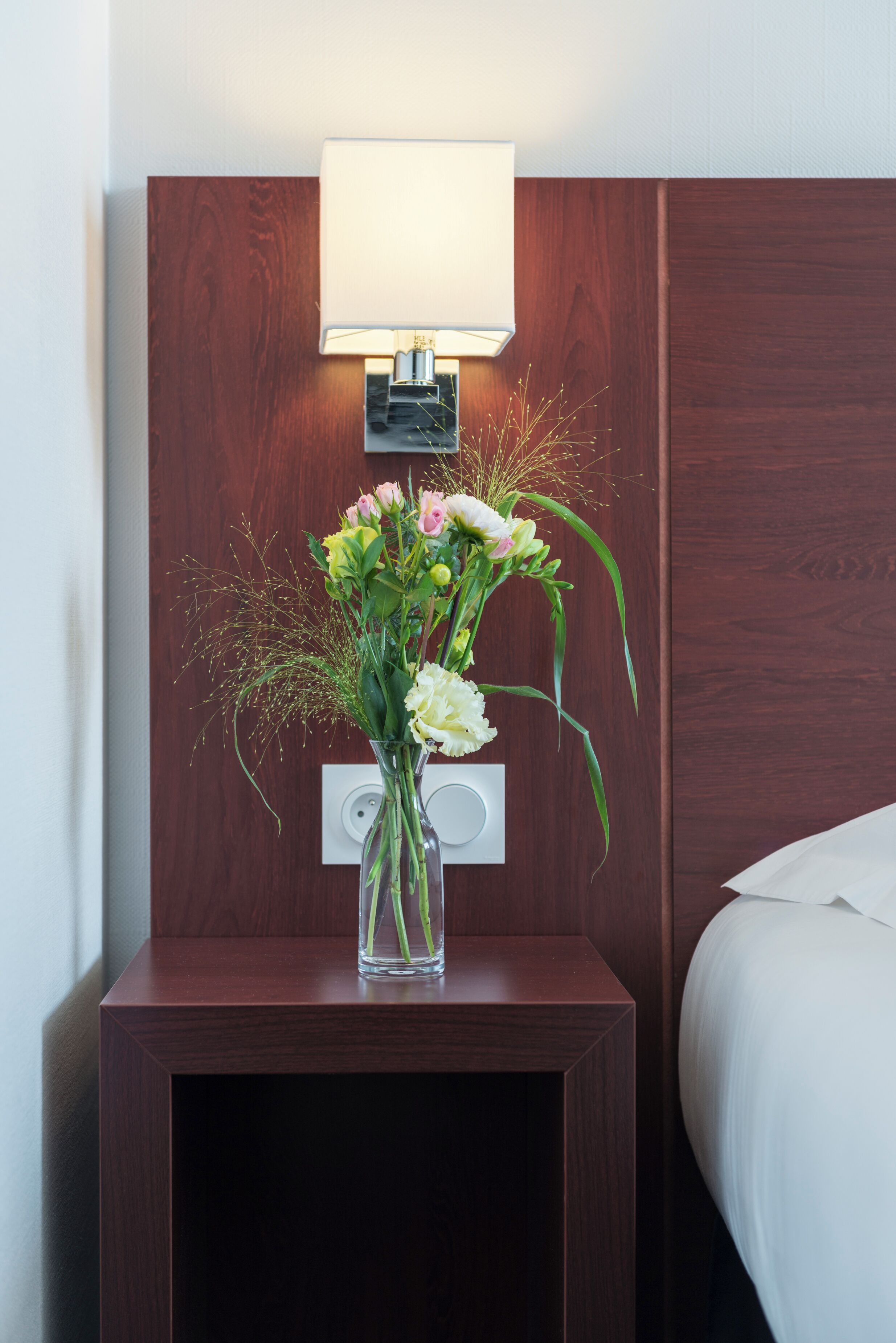 A wooden bedside table with a clear vase of flowers and a wall-mounted lamp.