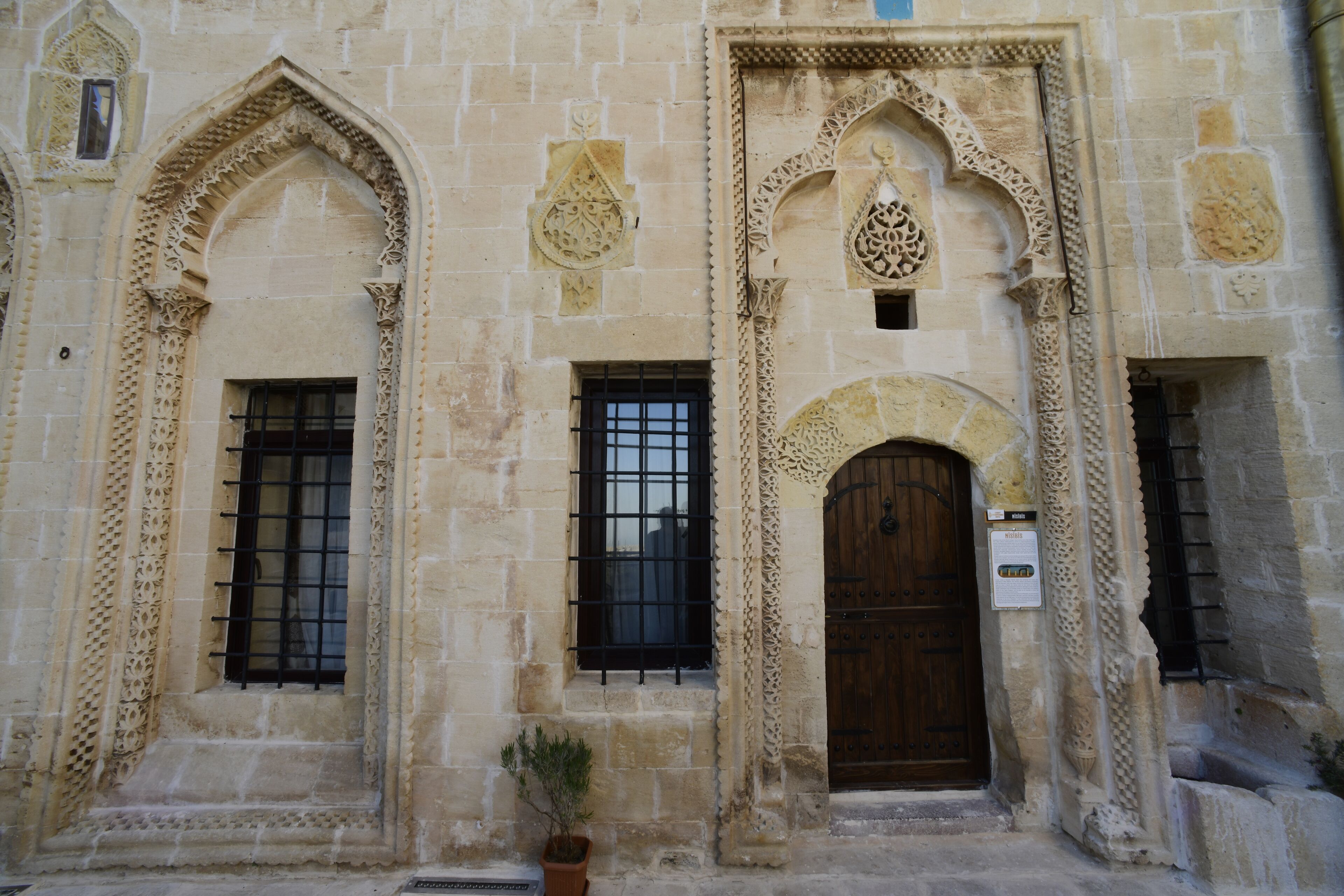 A building facade with arched windows and a wooden door, featuring intricate stone carvings and a small potted plant.