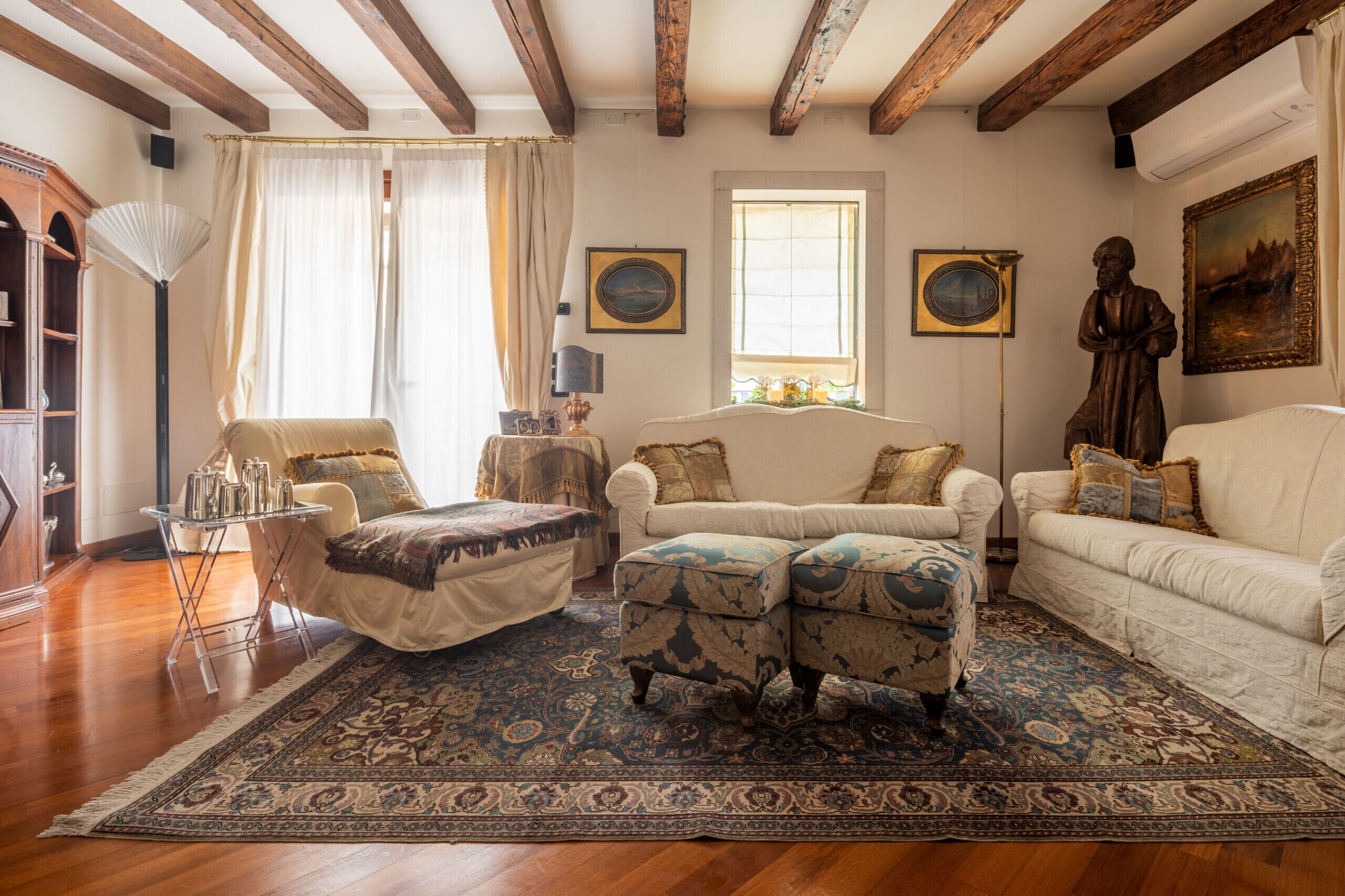 A living room with a sofa, ottoman, and armchair, a patterned rug, wooden beams on the ceiling, and a window with curtains.