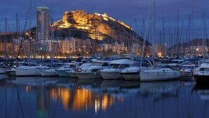 Un port de plaisance avec des bateaux amarrés, un bâtiment imposant illuminé, et une surface d’eau calme qui reflète les lumières.