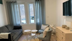 A modern living room with a grey sofa, a small armchair, two round tables, a wooden dresser, and a large window with curtains.