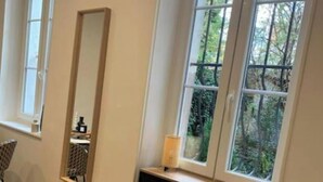 A room with a wooden floor, a black desk with a chair, a window with a view of greenery, and a mirror on the wall.