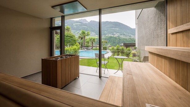 A modern room with a large window, a wooden bench, a small table, and a view of a pool and mountains. - Apartment "Wohnung 2" with Mountain View, Heated Pool & Wi-Fi (Riffiano)