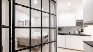 Une chambre moderne comprenant un lit, un bureau blanc, un coin cuisine avec un four à micro-ondes et une grande fenêtre aux montants noirs.