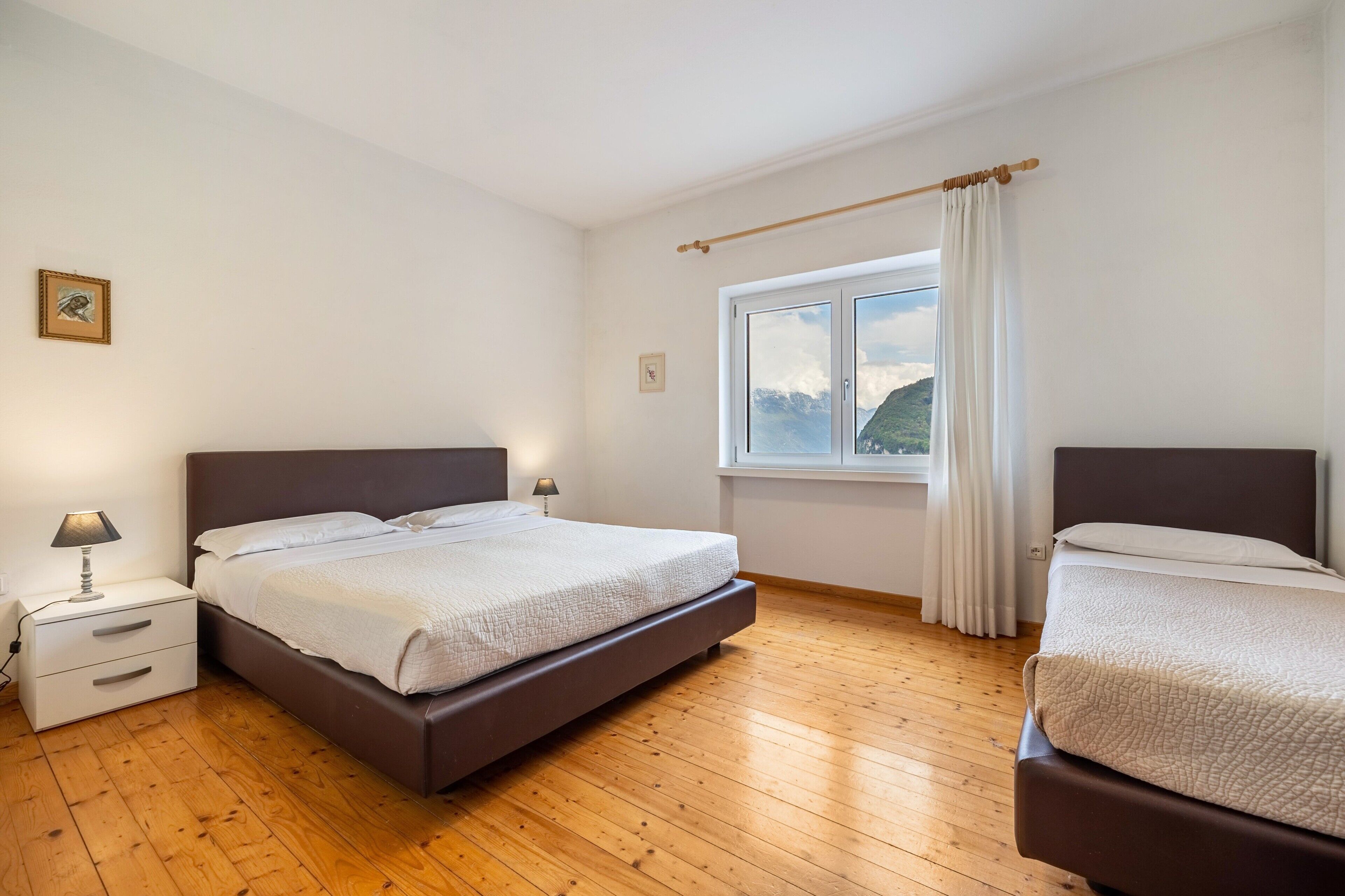 Una camera da letto con due letti, un comodino, una lampada, una finestra con tende e un quadro appeso al muro.