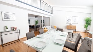 Een moderne eetruimte met een lange tafel gedekt voor zes personen, omringd door grijze gestoffeerde stoelen. Een zwart metalen console tafel met een cactus en boeken staat links. De kamer heeft een hoge plafond met een balkonhek, en er hangen ingelijste kunstwerken aan de muren.