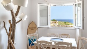 A dining area with a table and chairs, a wooden surfboard decoration, and a view of the sea through an open window. - Kalliston, Romantic Villa with Amazing Sea View (Naxos)