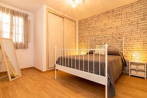 A bedroom with a metal bed frame, a brick wall, a wooden floor, a wardrobe, and a bedside table with a lamp.