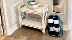 Esquina de una habitación con un radiador blanco, una pequeña mesa blanca con una cesta de objetos, un espejo y una cesta de mimbre con ropa doblada.