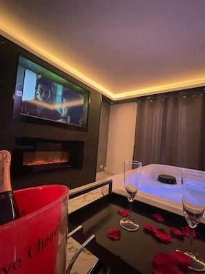 Een kamer met een aan de muur bevestigde televisie, een open haard, een tafel versierd met rozenblaadjes, en een rode emmer met de tekst 'Cliche' erop.