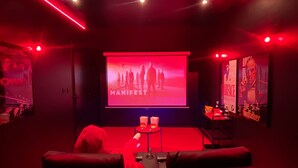 Une pièce sombre avec un grand écran diffusant l’image de « MANIFEST », un éclairage d’ambiance rouge et un espace détente avec une petite table et des collations.