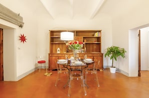 Un coin repas avec une table ronde en verre, des chaises transparentes, une plante en pot et un meuble en bois contenant des livres.