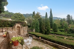 Une terrasse de jardin avec des tables et des chaises en pierre, une arche en pierre et une vue sur un paysage aménagé, avec des arbres et une colline au loin.