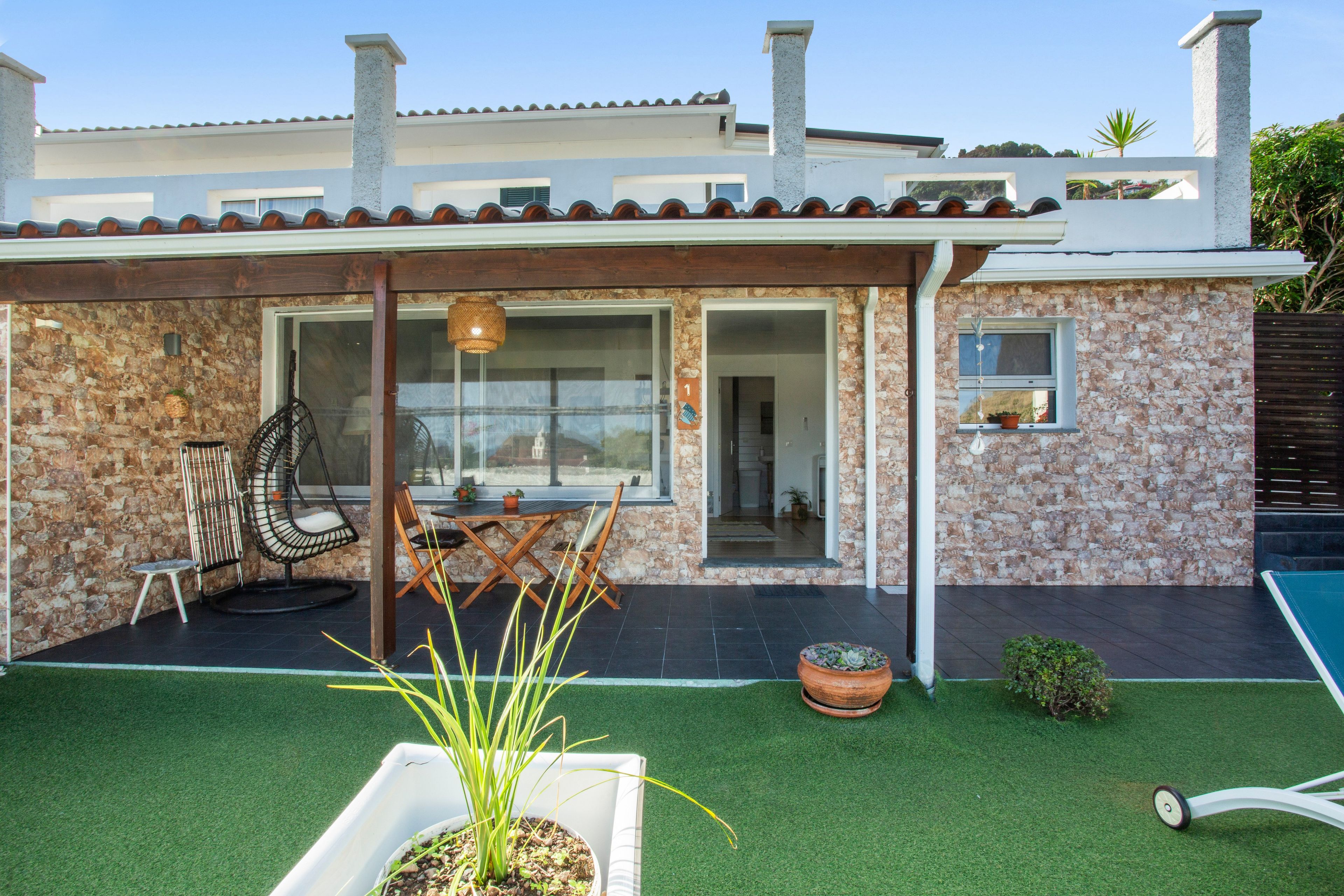 A house with a stone facade, a patio with a table and chairs, and a small garden area with artificial grass and potted plants.
