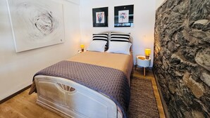 A bedroom with a bed, two bedside tables, a wall-mounted artwork, and a stone wall. - Studio Matilde & Joaquim – Panoramic Sea View (Porto da Cruz)