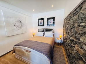 Un dormitorio con una cama, dos mesitas de noche, una pared decorada con obras de arte y una pared de piedra.