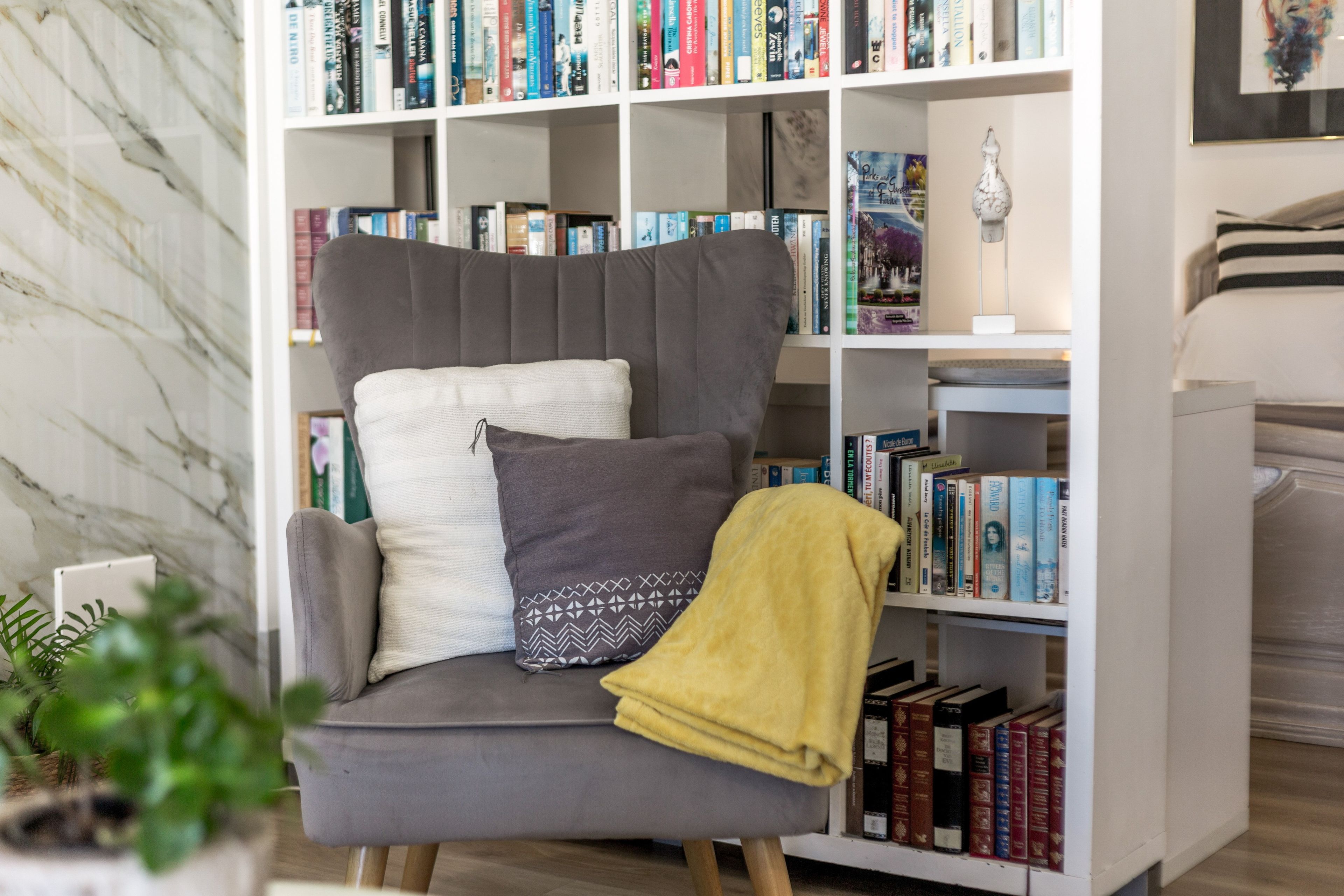 Un fauteuil confortable pour lire, une couverture, une bibliothèque remplie de divers ouvrages et un mur en marbre.