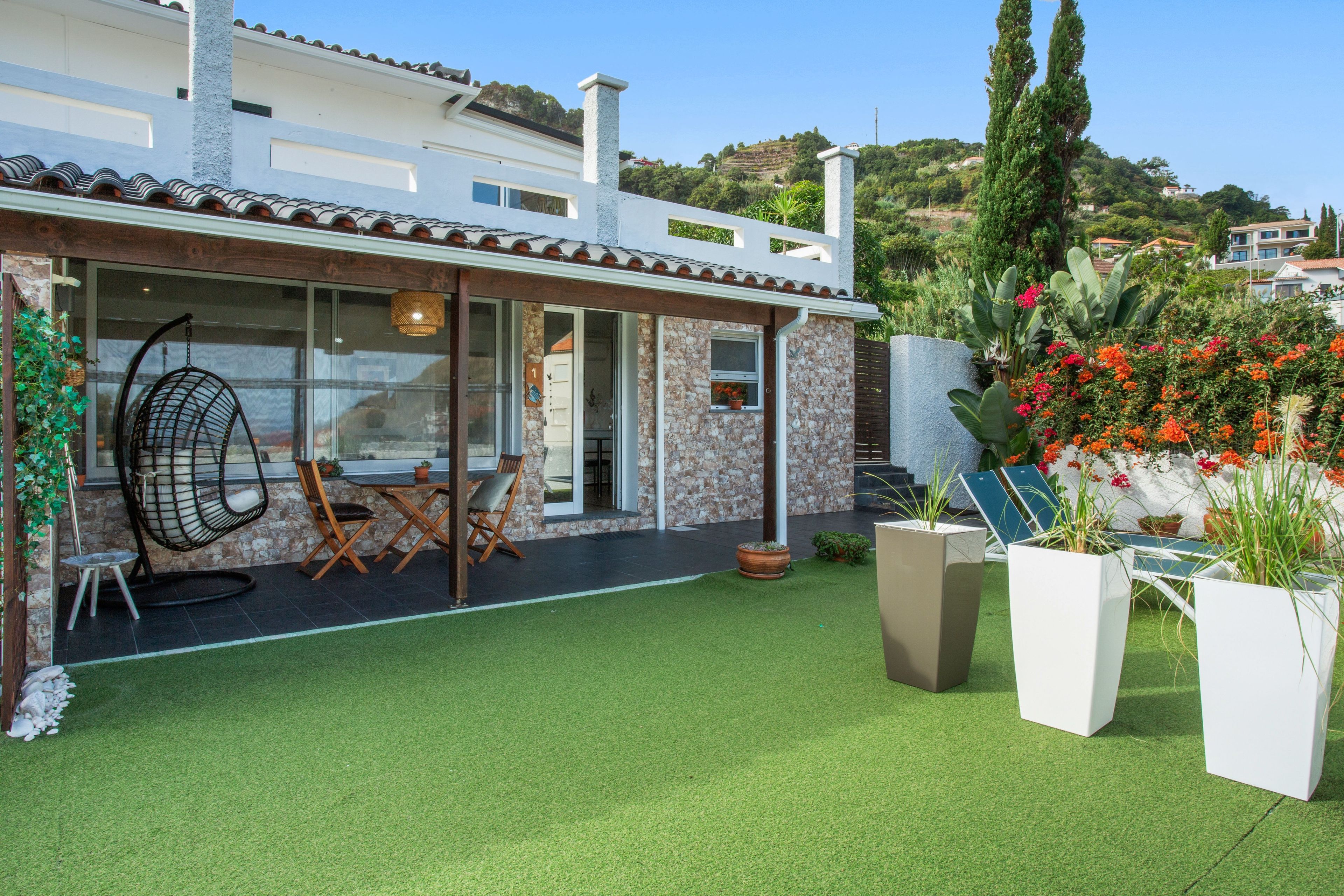 Une maison moderne dotée d’un patio, d’un coin repas avec des chaises et une table, et d’un jardin agrémenté de plantes en pot et de fleurs.