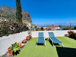 A terrace with two lounge chairs, potted plants, and a view of a coastal town and mountains. - Studio Matilde and Joaquim, panoramic sea view (Porto da Cruz)