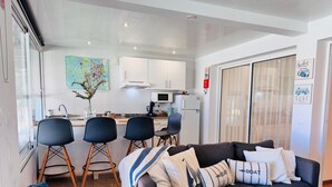 Ein modernes Wohnzimmer mit grauem Sofa, einem Essbereich mit vier Stühlen, einer Küche mit weißen Schränken und einem großen Fenster.