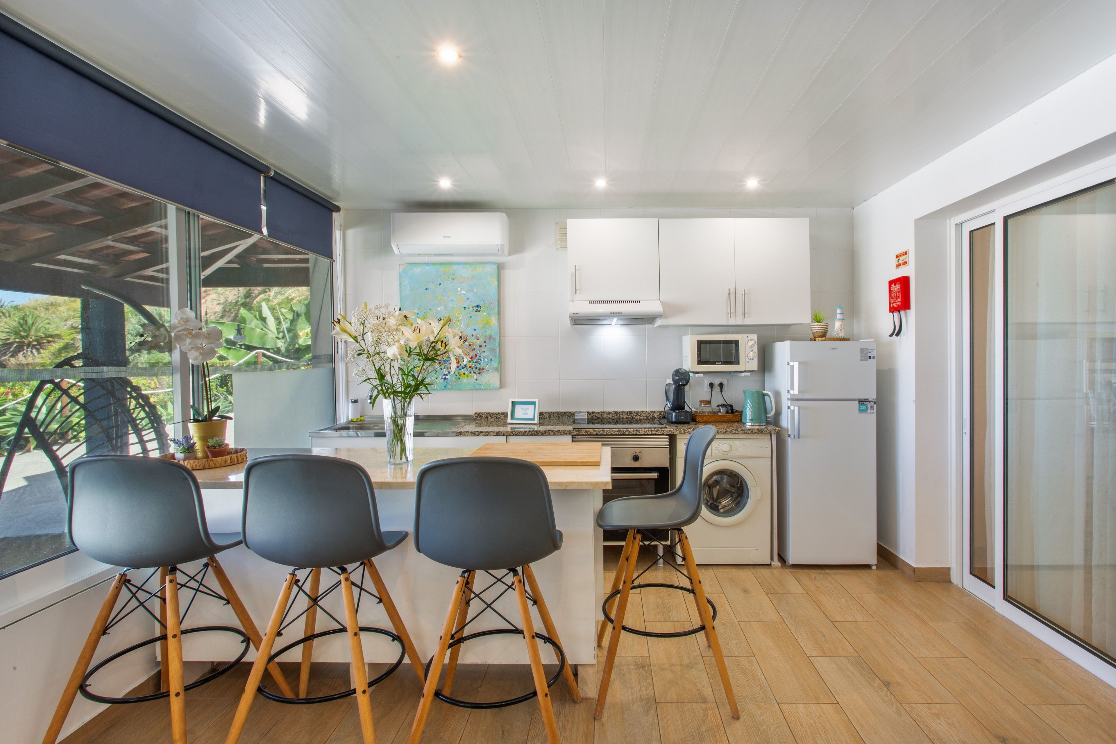 A modern kitchen with a central island, bar stools, a microwave, a washing machine, and a large window with a view of greenery.