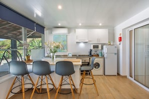 A modern kitchen with a central island, bar stools, a microwave, a washing machine, and a large window with a view of greenery.