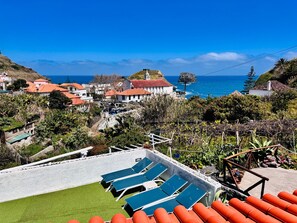 A coastal view with a pool area, lounge chairs, and a building with red roofs. - Villa Matilde e Joaquim, Sea View and Nature (Porto da Cruz)