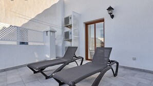 Zwei Liegestühle auf einer gefliesten Terrasse mit einer weißen Wand, einem Fenster und einer Klimaanlage.