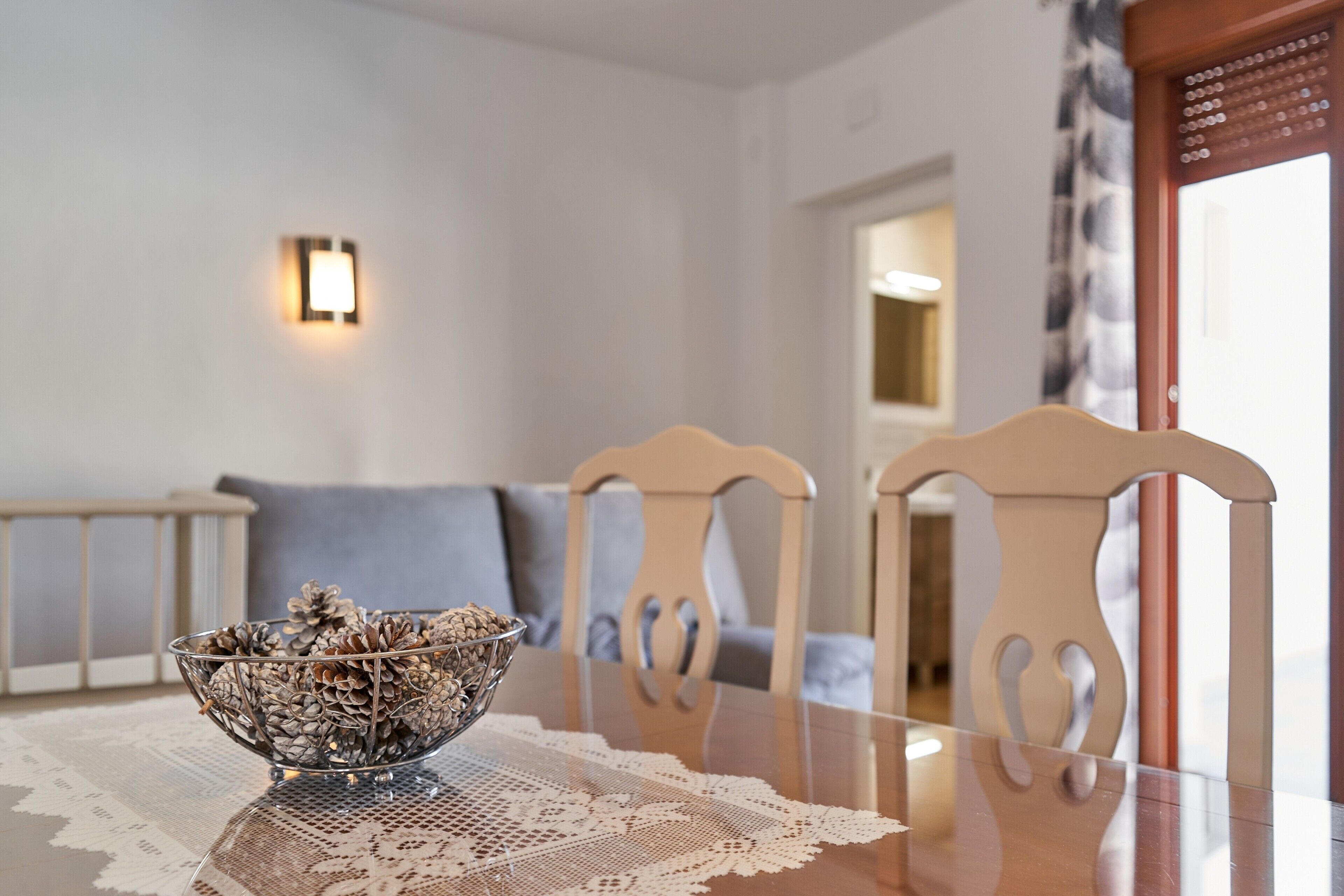 Una mesa de comedor con un cuenco de vidrio transparente que contiene piñas, un mantel de encaje y dos sillas de madera con acabado en tono claro.