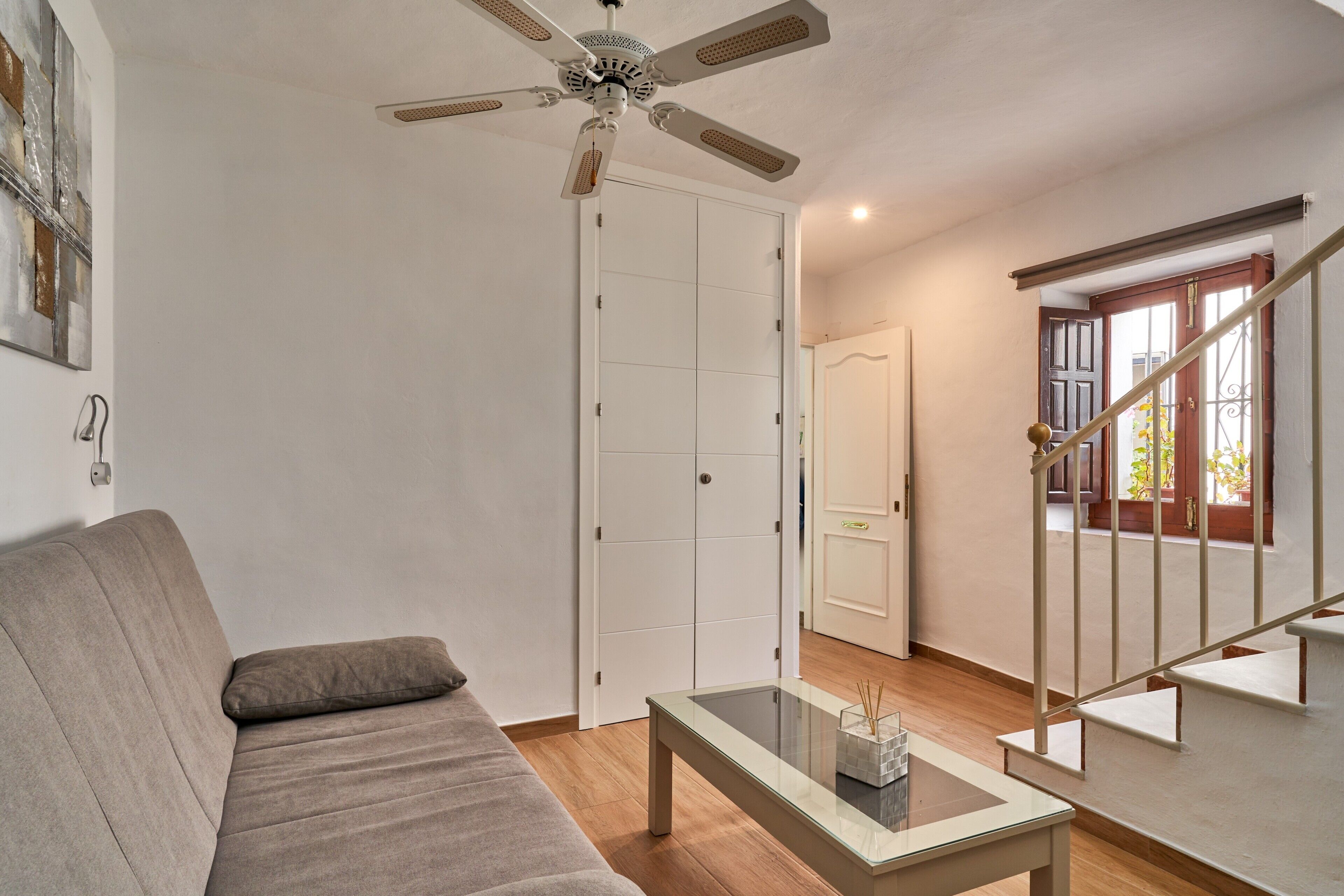 Uma sala de estar com sofá, mesa de centro de vidro, ventilador de teto, escada e porta que dá para outro cômodo.