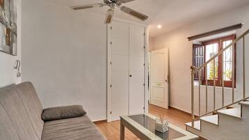 Uma sala de estar com sofá, mesa de centro de vidro, ventilador de teto, escada e porta que dá para outro cômodo.