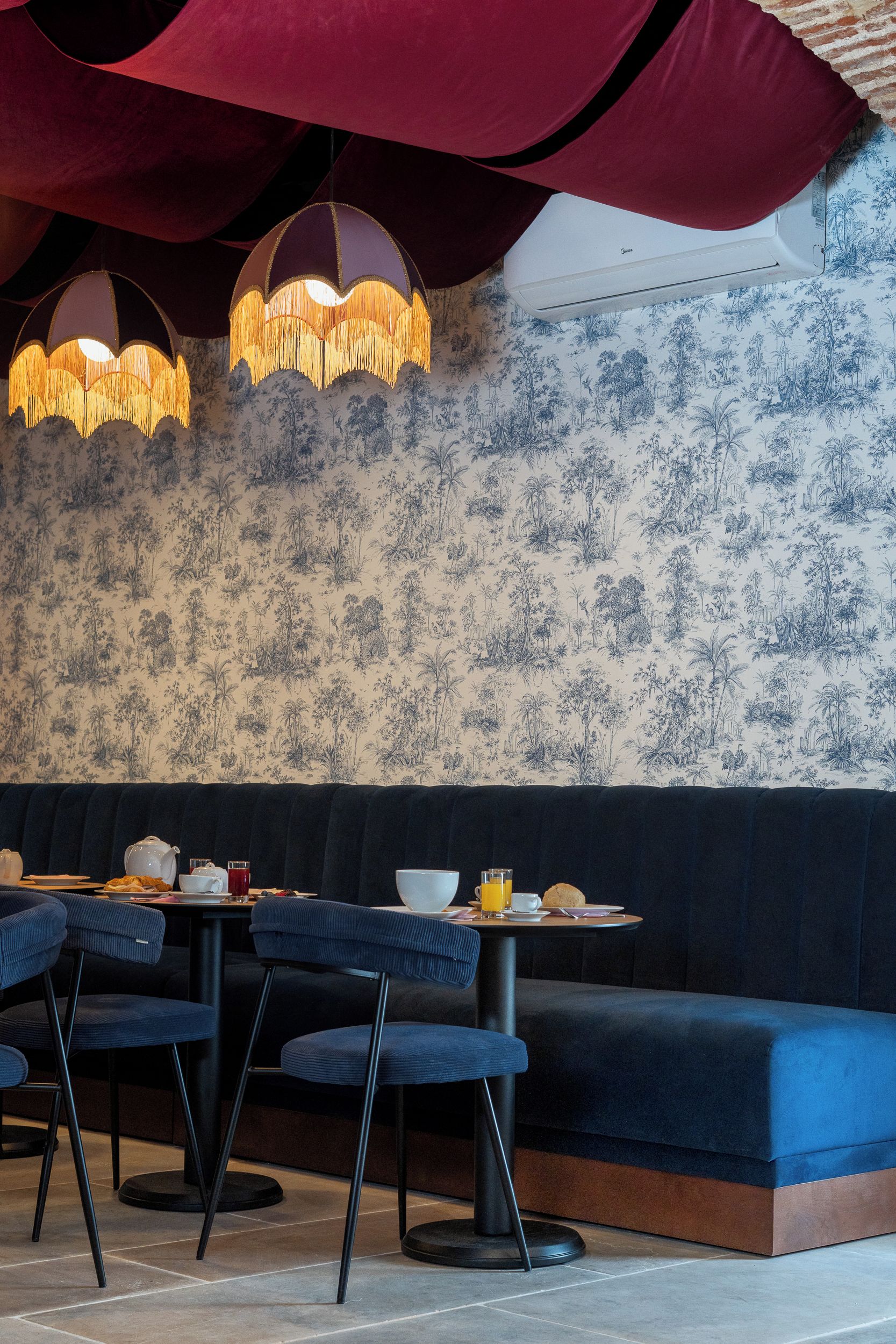 L’intérieur d’un restaurant présente un banc capitonné de couleur bleu foncé, une table ronde avec des chaises et un papier peint à motifs.
