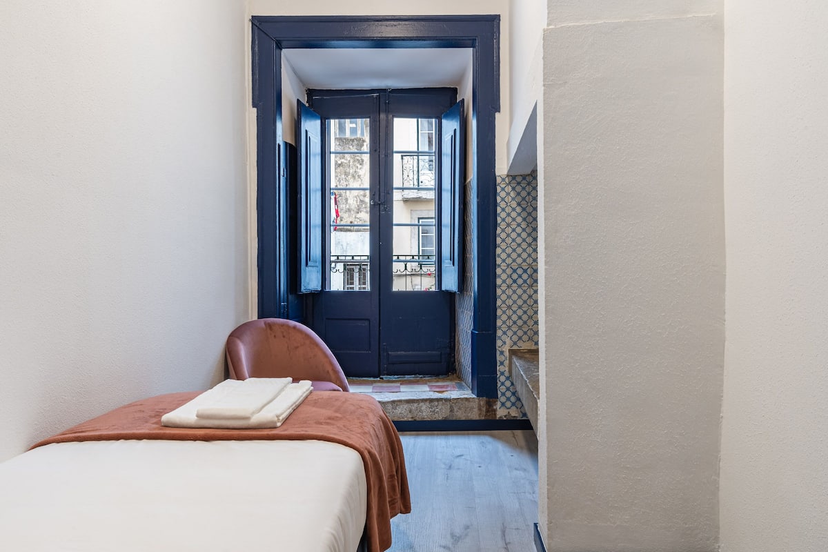Een smalle gang die leidt naar een donkerblauwe deur met een klein balkon. Een bed met een witte matras en bruine deken is aan de linkerkant te zien.