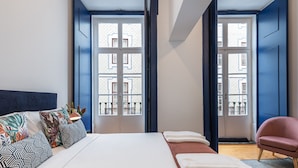 A bedroom with a large bed, two windows, a chair, and a view of a balcony. - Succeed Terreiro do Paço Suites (Lisbon)