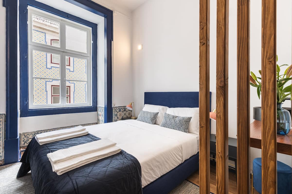 Een netjes opgemaakt bed met een blauw hoofdeinde, een raam met een decoratief patroon en een houten roomdivider.