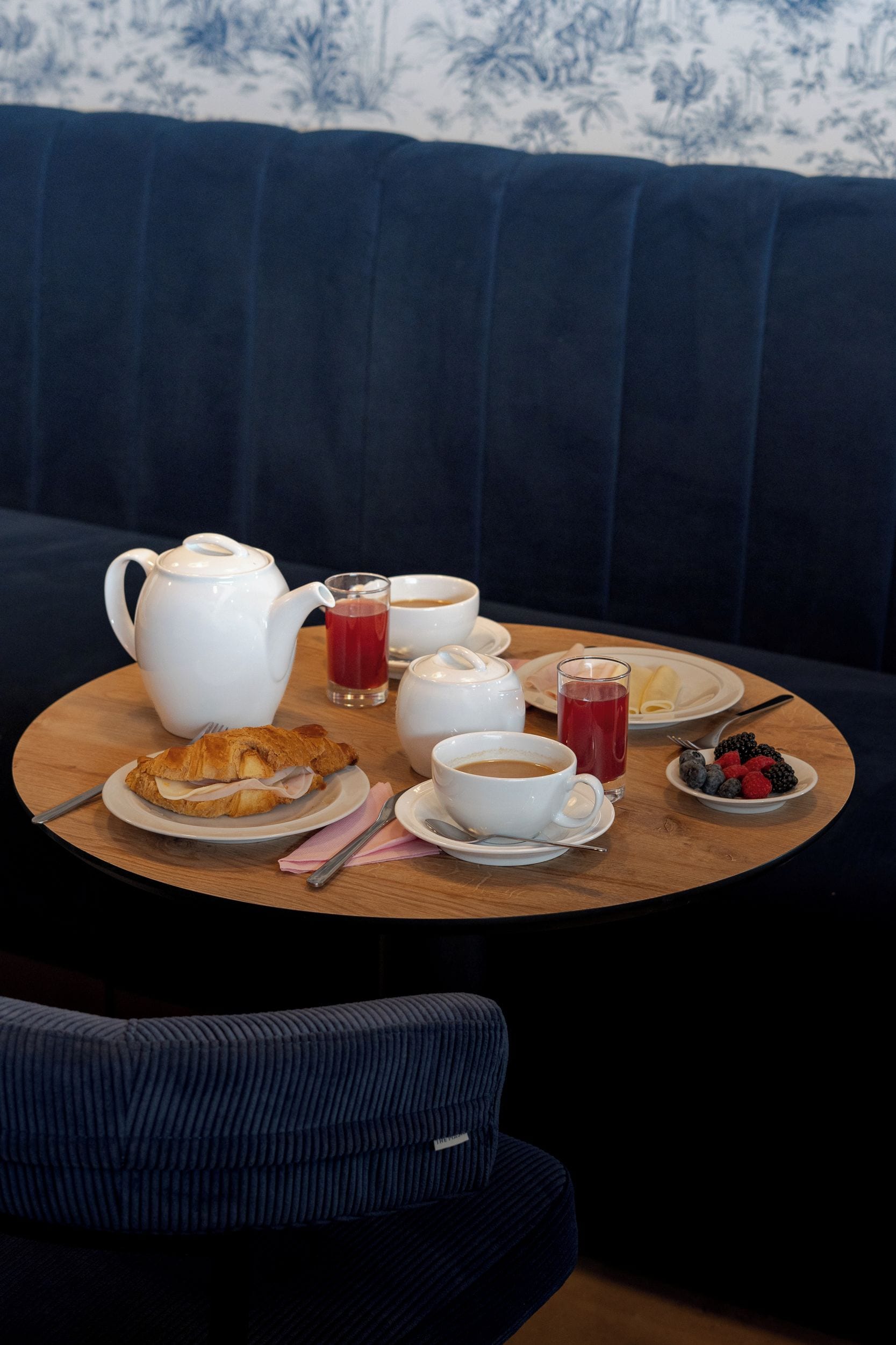 A round wooden table with a tea set, including a teapot, cups, and saucers, accompanied by a croissant, a glass of red beverage, and a plate of berries.