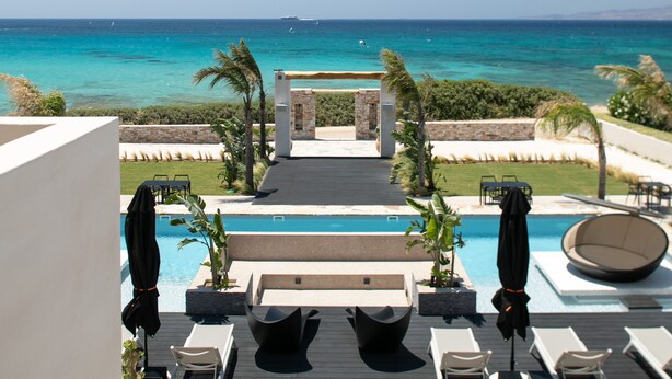 Zona de piscina con sillones reclinables y sombrillas, con vistas al mar azul y transparente.