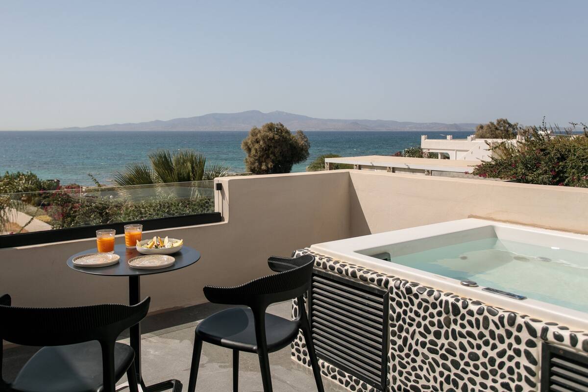 A balcony with a table, two chairs, a hot tub, and a view of the sea and mountains.