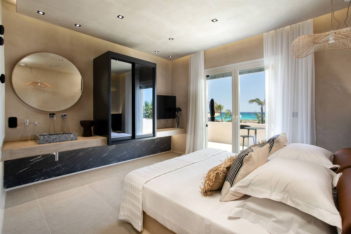 A modern bedroom with a large bed, a built-in TV, a bathroom vanity, and a balcony with a view of the ocean.