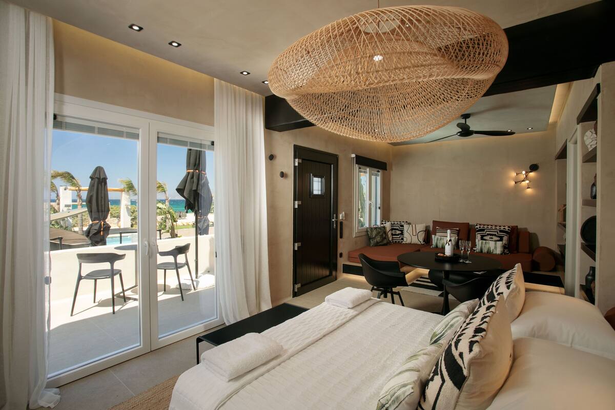 A modern hotel room with a large bed, a dining area, and a balcony with a pool view.