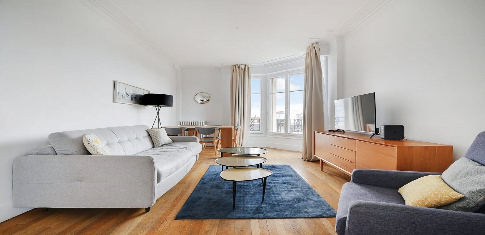 Un salon moderne avec un canapé gris, une table basse, un téléviseur posé sur un meuble en bois, et un coin repas à l’arrière-plan.