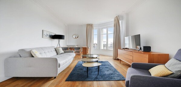 A modern living room with a grey sofa, a coffee table, a TV on a wooden cabinet, and a dining area in the background.