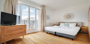 A bedroom with a large window, a bed, a dresser, and a television.